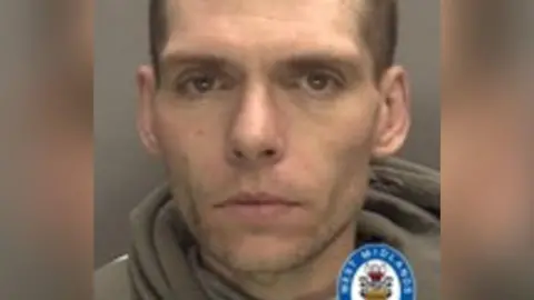 A mugshot of a man in a grey hoodie. He has a buzz cut and is staring into the camera.