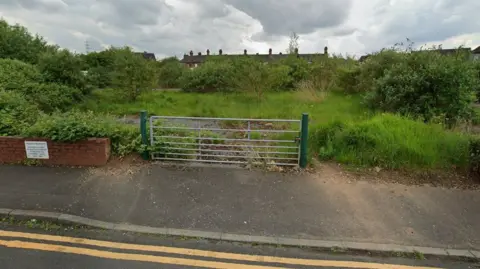 Google A patch of rough ground with grass and shrubs covering a hard-standing base. A metal gate and double yellow lines front the site, and the roofs of a row of terraced houses are visible behind.