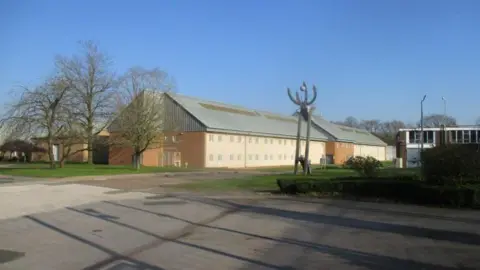 HMIP The exterior of several mid-20th century prison buildings with trees and grass surrounding them. A modern sculpture is visible in front of the buildings.