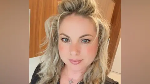 A woman with long, wavy blonde hair. She is wearing a black V-neck T-shirt and has a tattoo of a sparkle at the top of her chest.