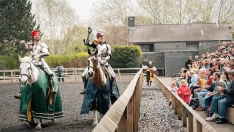 Royal Armouries Two men in armour mounted on horses in a recreated tiltyard setting with a large seated audience watching