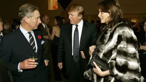 Getty Images Charles holds a drink while speaking to the Trumps. Melania wears a wool coat and clutches a purse