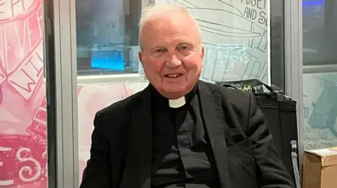 Shows a man with grey hair in a black suit. He is sitting down.