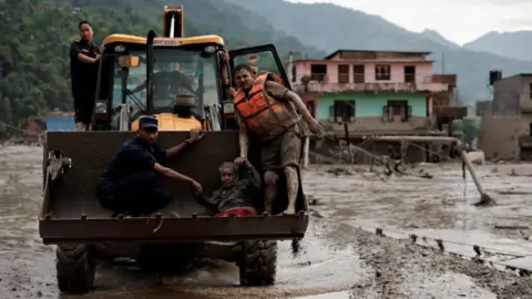 Reuters Rescuers use a digger truck to carry an elderly woman to safety. Buildings swamped by mud can be seen in the background