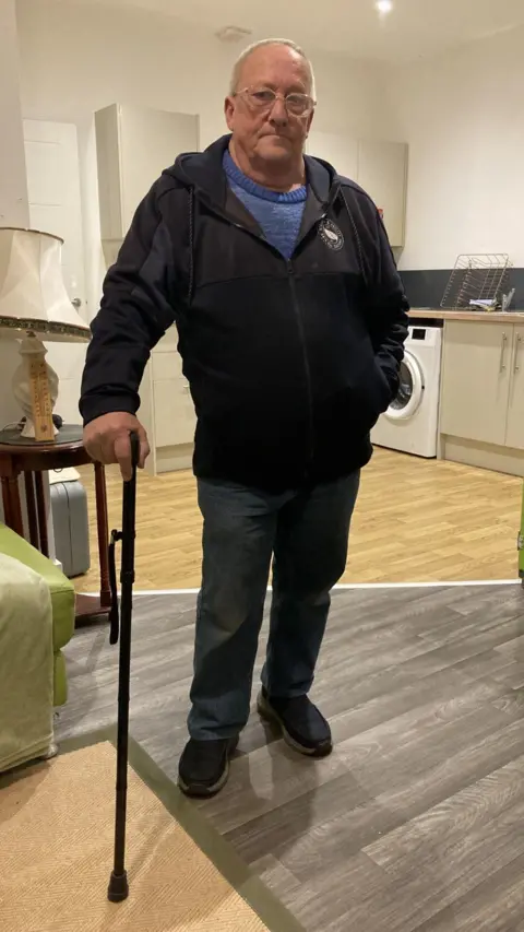 A man in his 60s is stood in an open plan kitchen/living room. He has short grey hair and wears glasses. One hand is leaning on a walking stick. He is wearing blue jeans, a blue T-shirt and a navy blue zipped hoodie.