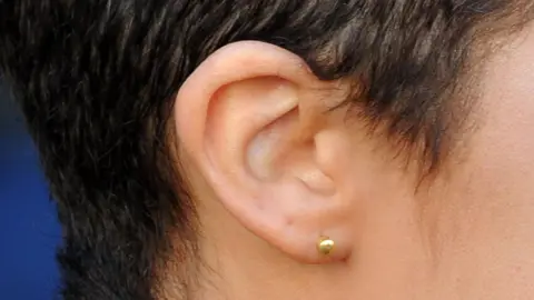 PA Media A close-up of a woman's ear. She ha short dark hair and a small gold stud earing.
