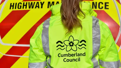 Cumberland Council A woman wearing a high-vis jacket with the words 'Cumberland Council' on the back faces the rear of a highway maintenance van