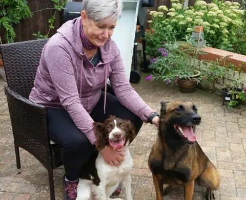 Humberside Police Sharon sits in a chair on the garden patio. She wears a dusty pick hoodie and leans forward with her hands on both dogs. Quinta looks like a alsation cross, and the other dog is some kind of spaniel.