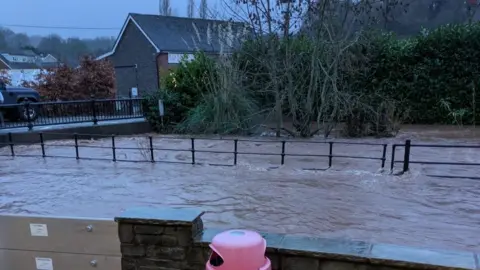 A street in Ewyas Harold is flooded with murky water. A building and road are visible in the background and there is the top of a red bin next to a wall in the foreground.