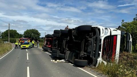 Overturned lorry at the side of a road, with police and firefighters