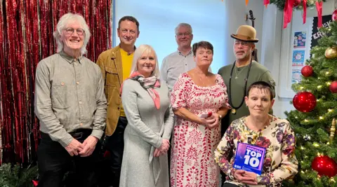 A man with long grey hair, grey shirt and glasses, a man with brown hair and a yellow top, a woman with grey hair and a long grey dress, a man with grey hair and a white shirt, a woman with dark hair and a red and white patterned dress, a man with a green tshirt and brown bowler hat and a woman with short dark hair and a floral patterned dress, holding a blue and pink trophy