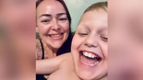 Family photograph Close-up of Gemma with long brown hair pictured sitting on a dark green sofa with her son, William, nine, who has blond hair. They are both laughing.