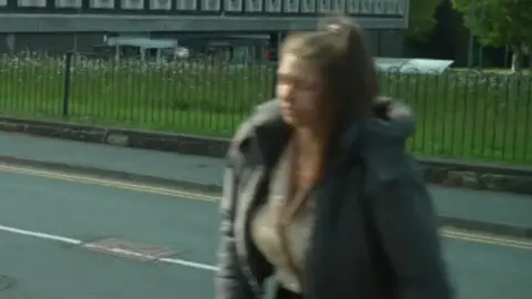 A blurred photo of a woman walking across a road. She has her brown long hair tied up in a ponytail. she is wearing a grey coat.