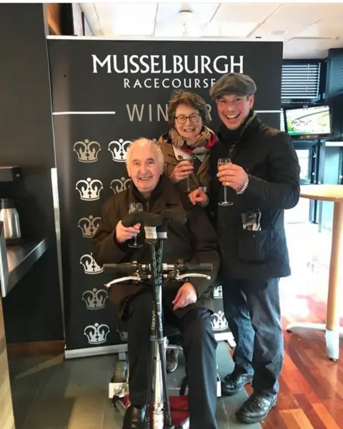 James Ewart Racing Three people, one sitting down and two standing up raise glasses of champagne to celebrate a winning horse at Musselburgh Racecourse