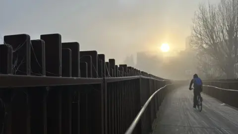 A young person rides their bike down a wooden walkway. On the left is a wooden fence where you can see the spiders webs laced with frost. In the distance the sun is just beginning to rise.