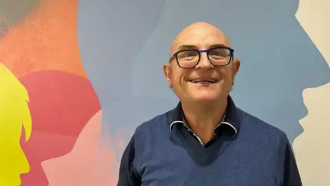 Ollie Conopo/BBC A man looking at the camera, smiling, with glasses, a dark blue shirt and blue jumper. He is standing in front of a colourful wall.