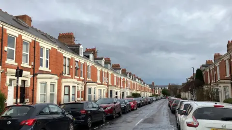 A street full of Victorian houses most with bay windows. They are redbrick with white windows and lots of chimneys. Rows of cars are parked outside.