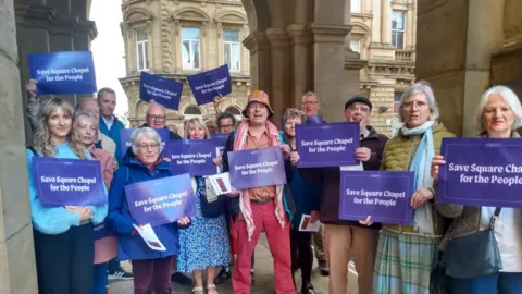 14 people standing with purple placards that read Save Square Chapel for the People