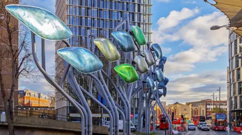 Getty Images Large curved metallic leaf sculptures in Stratford, east London, with high‑rise buildings and red double‑decker buses in the background.
