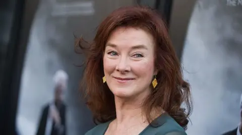 Valerie Mahaffey is pictured in front of a grey background. She has red hair and is wearing a green dress and gold earrings. She has blue eyes.