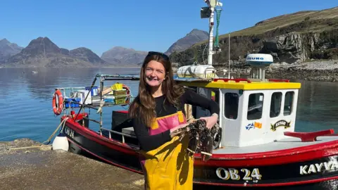 James Robertson Brooke Lamond holds up a lobster in front of a small creel fishing boat. She wears a long-sleeve black top with yellow fishing overalls on top. It is a sunny day and you can see the water behind her and hills in the distance.