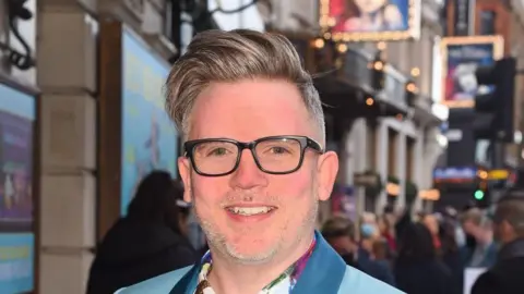 Tom MacRae smiles at the camera outside a theatre. He is wearing a blue jacket and back-rimmed glasses. His hair is styled in a quiff. 