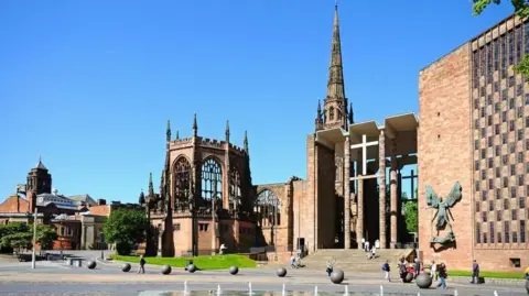 Getty Images Coventry's old cathedral ruins seen next to the new 20th Century building. There is a clear, blue sky and people are walking around the cathedral precincts. There are decorative globes and a water feature is visible.