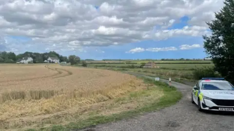BBC/Gemma Dawson A photos of a countryside road leading up to the shooting centre. A police car is parked facing the camera in the middle of the road. On the left is fields of hay and on the right are grassy fields in the distance. The sky is blue with clouds. 