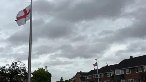 A St George's flag on a lamppost on the left-hand side of the road, and a Union jack flag on a lamppost on the right-hand side of the road. There are houses on either side.