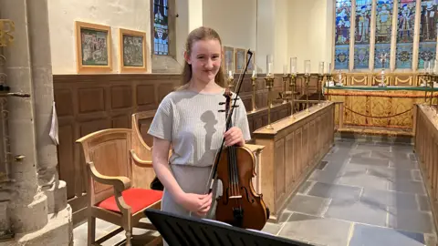 A teenage girl playing a viola. The background is out of focus but pictures in gold frames can be made out hanging on the walls
