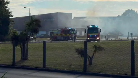 Manx Radio Fire engines outside the fire station. The image is taken through a fence surrounding the site. The smoke has thinned out. Some fire officers can be seen in front of their vehicles. 