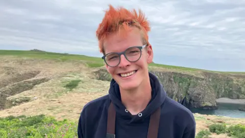 A man looks at the camera and smiles. He is stood on Skomer island and you can see the land and a bit of the sea behind him. He has a blue hoody on and is wearing glasses. He smiles at the camera and ginger hair which is spiky and blowing in the wind.
