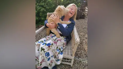 Contributed A woman is sitting in a chair on a gravel drive wearing a flowery skirt and blue jacket. A dog is sitting on her lap, trying to lick her face and she is pulling away laughing 