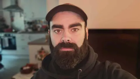 Chris Cullen A picture of Chris close up in his apartment. He has brown eyes and dark beard and brows and a nose ring. 