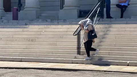 Steps of crown court and woman walking down last steps in black jeans and white trainers with a black shoulder bag and loose beige top. She is holding a grey jumper over head to cover her face and hair.
