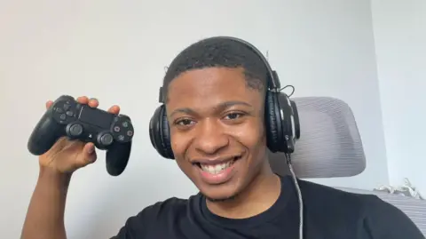 Dienne Kawende Dienne, a man with short hair, smiles into the camera wearing big gaming headphones. He's wearing a black t-shirt sat on a grey chair with a head rest. He's holding up a black Playstation 2 controller.
