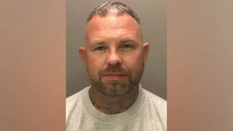 A police custody photograph of Michael Watling. He is wearing a grey jumper. Watling has short, greying hair and a short beard. His neck is also covered in tattoos.