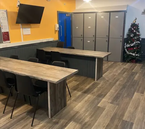 Two breakfast benches with a wooden top, surrounded by stools on a wooden floor, grey lockers are visible against a wall with a christmas tree beside them, the walls are bright yellow with a television hanging 