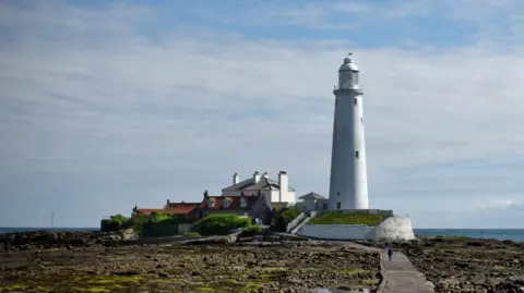 St Mary's Lighthouse in Whitley Bat. The causeway is revealed because the tide is out. The lighthouse is white, slim and has a number of windows running up the building. A house stands next to it.