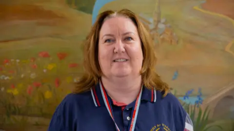 Julie Wadham. She has shoulder length brunette hair and is wearing a navy blue Help for Heroes polo shirt with white and red stripes on the collar. She is looking directly at the camera and smiling. 