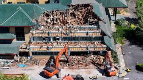 Eddie Mitchell An aerial image of demolition works on a square building with a dark green roof. The building is surrounded by cranes and has a chunk taken out of its side. the roof has a number of triangular layers.