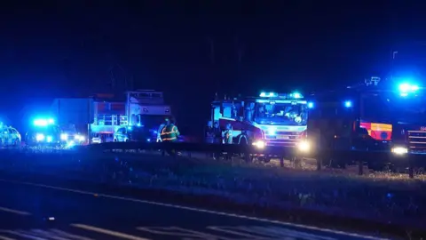 Emergency service vehicles, including fire engines, police vehicles and lorries, are stopped on the A27 at night. Blue emergency lights illuminate the scene while several people stand near the central reservation.