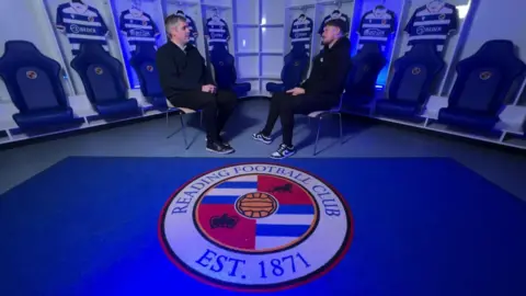 Two men, both wearing black trousers and hoodies, sitting on chairs facing each other in mid-conversation. In front of them is a carpet the the Reading FC logo on it.