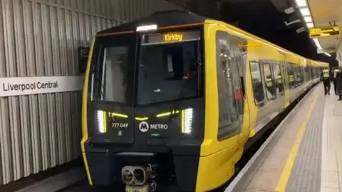 A yellow train with the destination Kirkby at Liverpool Central station platform. Staff in hi-vis vests and a passenger are seen in the background.