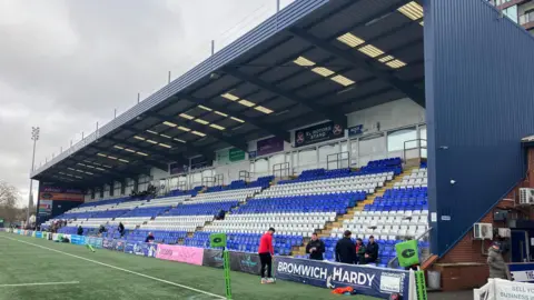 The main stand at the Butts Park Arena features a blue corrugated roof and side panels with banks of blue and white seating.