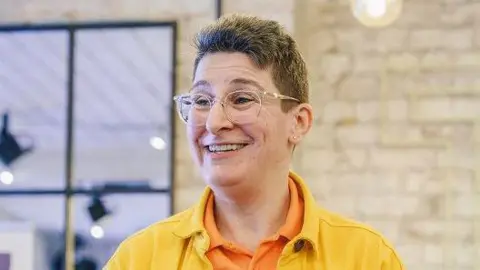 PA Media Sunny Turner, who wears glasses, smiles as she talks at an event. She wears a yellow shirt.