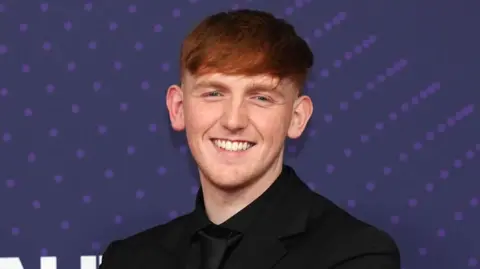PA Angry Ginge stands smiling in an all black suit against a purple backdrop