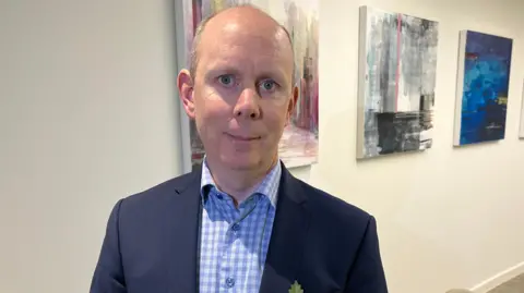 Jozef Hall/BBC Diarmuid O'Brien wears a blue checked shirt with a navy jacket and red poppy and is standing up with painting behind him.