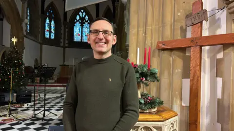 A man in a green jumper standing in a church in front of a wooden cross