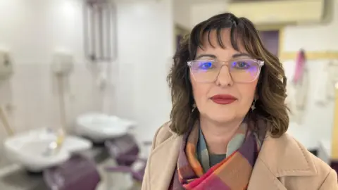 Elizabeth Baines/BBC A woman with brown hair and clear glasses stands in front of a hair dressers. She wears a beige coat and colourful scarf.